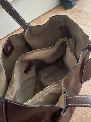 Brun shoppingväska med innerficka - Fin brun shoppingväska i nyskick, aldrig använd. Pris 250kr. Har en innerficka som man kan ta av och på, samt en knapplösning i mitten av väskan för att kunna stänga den. Stilren och rymlig design som funkar till mycket.