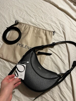 Svart axelväska Polène Cyme Mini - Säljer en svart Polène Cyme Mini axelväska i stilren halvmåneform. Väskan är gjord i strukturerat skinn och har guldfärgade detaljer samt justerbar axelrem. Kommer med dustbag och extra rem. Perfekt för dig som gillar minimalistisk och modern design.