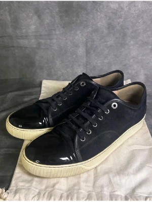 Lanvin Cap toe shoe - Säljer ett par svarta Lanvins sneakers med snygg lackad tå och vit sula. Skorna har snörning och är tillverkade i mocka och syntetmaterial. Den rundade tån och kontrasten mellan matt och blankt gör dem riktigt stilrena och unika. 43 passar 44