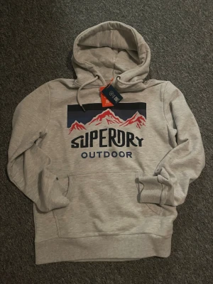 Superdry Hoddie - Grå superdry hoddie med tryck. Helt ny med prislapp. Storlek M. Hör gärna av dig vid minsta fundering!!