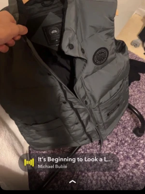 Grå dunväst från Canada Goose - Säljer en grå dunväst från Canada Goose med klassisk logga på bröstet och tydlig märkning på fickan. Västen har praktiska fickor, knappar och dragkedja framtill. Perfekt för lager-på-lager och snygg till streetwear. Stilsäker och funktionell design.