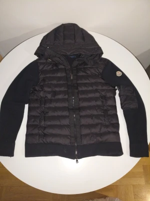 Svart pufferjacka från Moncler - Svart pufferjacka från Moncler med stickade ärmar och huva. Jackan har quiltad front, dragkedja och Moncler-logga på ena ärmen. Perfekt för dig som vill ha en snygg och varm jacka med coola detaljer.