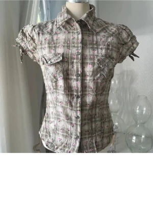 Kortärmad rutig skjorta med blommor - Kortärmad skjorta i beige och grå toner med rutigt mönster och små rosa blommor. Skjortan har tryckknappar, två bröstfickor och knytband på ärmarna. Materialet känns lätt och luftig, perfekt för varmare dagar. Storlek 38 men lite mindre i storlek, som S