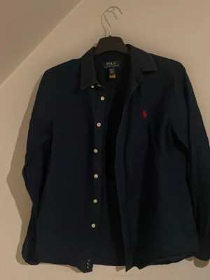 Mörkblå skjorta från Polo Ralph Lauren - Snygg mörkblå skjorta från Polo Ralph Lauren med klassisk krage och vita knappar framtill. Skjortan har lång ärm och den ikoniska röda Polo-loggan broderad på bröstet. Tillverkad i mjukt bomullstyg som känns skönt mot huden.