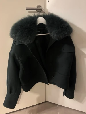 Svart pälsjacka från Meotine - Säljer en svart pälsjacka från Meotine med stor fluffig krage i päls. Jackan är croppad med breda muddar och har knappar framtill samt fickor på bröstet. Perfekt för dig som vill ha en statement piece med lyxig vibe.