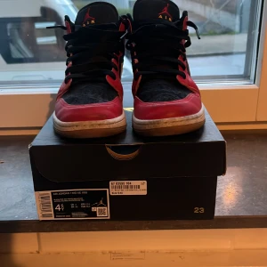 Nike Air Jordan 1 Mid SE svart/röd - Säljer ett par Nike Air Jordan 1 Mid SE i klassisk svart och röd färg med vita detaljer. Skorna har svart snörning, rund tå och den ikoniska swoosh-loggan på sidan. Ovandelen är i skinn och syntet, med en bekväm platt sula. Perfekta för dig som gillar streetstyle.