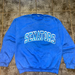 Blå college-tröja med SENATORS-tryck storlek M - Hösten är här och vad passar bättre än en klassisk crewneck? Den här blå tröjan har stort SENATORS-tryck i vitt och blått över bröstet. Rund hals och ribbade muddar gör den både bekväm och tidlös.  Perfekt om du gillar den sportiga, avslappnade looken och vill ha något varmt men ändå stilrent.