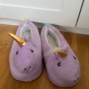 Lila enhörningstofflor med horn - Supergulliga lila tofflor i plysch med enhörningsansikte, guldigt horn och regnbågsfärgad man. Perfekta för att mysa runt hemma och ge en magisk känsla till vardagen. Mjuka och bekväma med rund tå och platt sula.