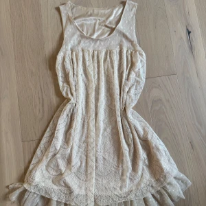 Beige spetsklänning med volang - Supersöt beige kortklänning med spetsmönster och transparent mesh upptill. Klänningen är ärmlös och har en volangkant nertill i prickig mesh som ger en extra touch. Perfekt för sommaren eller studenten.