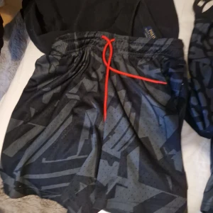 Jordan x PSG svarta meshshorts - Svarta meshshorts från Jordan x PSG med grått grafiskt mönster och röd snörning i midjan. Shortsen har elastisk midja och patch med logga på benet. Perfekta för sport eller chill, riktigt snygg streetstil.köpt från nike hemsidan gamla men knappt använda ny pris var runt 600 mitt pris 350 pris kan diskuteras vid snabbaffär