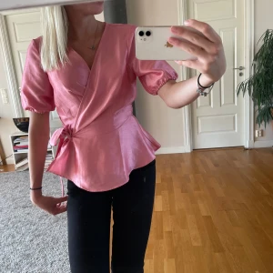 Rosa omlottblus med puffärm - Säljer en superfin rosa omlottblus med korta puffärmar och knyt i midjan. Blusen har en lätt glansig finish och markerad midja som ger en snygg siluett. Perfekt för dig som vill ha något extra till outfiten! Storlek m men passar mig jättebra som brukar ha xs/s💗