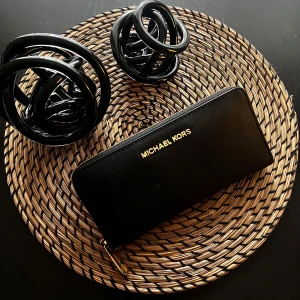 Plånbok från Michael Kors - Svart plånbok från Michael kors. Klassisk rektangulär form med logga i guld på framsidan. Den är fin och bra sicket.  Perfekt för att hålla koll på kort, cash och småprylar.