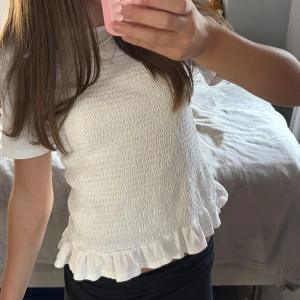 Vit smockad topp med volangkant - Supersöt vit topp med smockat liv och volangkant nertill. T-shirtrmar och rund halsringning ger en enkel men gullig look. Perfekt till jeans eller kjol för en fräsch stil. Materialet känns mjukt och stretchigt. Obs‼️sömmen har gått upp pyttelite på ryggen på ett ställe men inget som syns tydligt!🩷