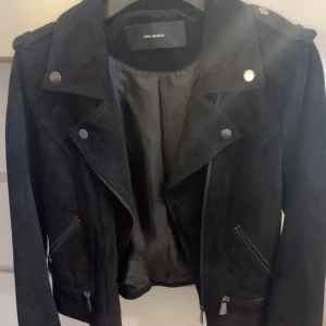 Svart mockajacka från Vero Moda - Snygg svart skinnjacka i mocka från Vero Moda med klassisk bikerstil. Jackan har silvriga dragkedjor, knappar och detaljer, samt breda slag och långa ärmar. Perfekt för dig som vill ha en edgy look.