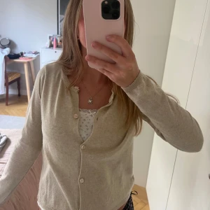 Beige kofta med knappar - Säljer en klassisk beige kofta med rund halsringning och knäppning framtill. Koftan har långa ärmar och ribbade muddar vid ärmslut och nederkant. Perfekt att slänga över en topp för en chill och stilren look.