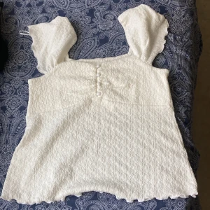 Vit blus med puffärmar från Feather & Bone - Supersöt vit blus från Feather & Bone med puffiga korta ärmar och vågig kant nertill. Toppen har en strukturerad, krinklad yta och dekorativa knappar framtill. Perfekt för dig som gillar romantisk och trendig stil. Aldrig haft på mig fick som present.