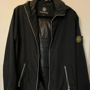Svart vindjacka från Stone Island - Svart vindjacka från Stone Island med ikonisk patch på ärmen. Jackan har dragkedja framtill, två fickor med dragkedja och justerbar nederkant med snörning. Tillverkad i polyester och har en clean, sportig look som passar perfekt till streetwear.