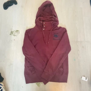 Vinröd hoodie från Polham med brodyr - Vinröd hoodie från Polham med broderad logga på bröstet. Tröjan har hög krage med tre knappar, dragsko i huvan och klassisk känguruficka. Mjuk bomullskänsla och långa ärmar, perfekt för kyliga dagar.