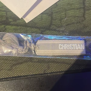 Beige broderat armband från Dior - Säljer ett snyggt beige armband från Dior med broderad text 'CHRISTIAN DIOR J'ADIOR' och dekorativa tofsar i ändarna. Armbandet är gjort i textil och har en stilren, neutral look som passar till många outfits.