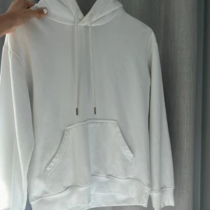 Vit hoodie med huva och ficka - Klassisk vit hoodie med huva och dragsnören. Tröjan har en stor magficka och långa ärmar. Perfekt för en chill och avslappnad stil. Enkel och stilren design som passar till det mesta.