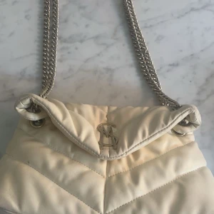 Vit quiltad axelväska Steve Madden - Snygg vit axelväska från Steve Madden med quiltad design och silverfärgad kedjerem. Väskan har ett stort fack, innerficka med dragkedja och logga framtill. Perfekt för dig som vill ha en trendig och stilren väska med plats för det viktigaste.