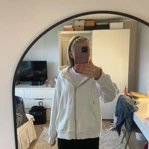 Vit hoodie med dragkedja  - Säljer en vit hoodie med dragkedja och huva. Tröjan har två stora fickor framtill, ribbade muddar och en avslappnad passform. Perfekt för dig som gillar en enkel och clean stil. Materialet känns mjukt och skönt mot huden.