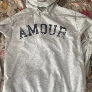 Ljusgrå hoodie från Gina Tricot - Ljusgrå hoodie från Gina Tricot med svart text 'AMOUR' framtill och en stjärna på ryggen. Tröjan har huva med snörning och är tillverkad i mjuk bomullsblandning. Perfekt för chill dagar och enkel att styla med jeans eller leggings.