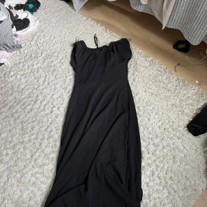 Svart långklänning med knyt - Svart långklänning med smala axelband och v-ringning. Klänningen har ett snyggt knyt framtill och lätt rynkade detaljer vid axlarna. Materialet är mjukt och faller fint längs kroppen, perfekt för dig som gillar en enkel men stilren look.