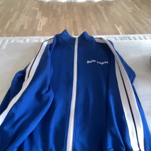 Blå track jacket från Palm Angels XL - Blå track jacket från Palm Angels med vita ränder längs ärmarna och vit dragkedja framtill. Jackan har hög krage och logga på bröstet. Tillverkad i bomull och polyester för en sportig vibe. Perfekt för dig som gillar streetwear och vill sticka ut.