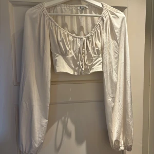 Vit croppad blus från Chiquelle - Supersöt vit croppad blus från Chiquelle med långa puffärmar, rynkad urringning och knyt framtill. Toppen har dekorativa knappar och är tillverkad i ett mjukt, lätt tyg som känns skönt mot huden. 