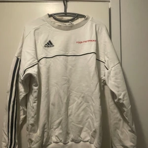 Vit Adidas x Gosha Rubchinskiy tröja - Vit crewneck från Adidas och Gosha Rubchinskiy med broderad rysk text i rött och klassiska svarta stripes längs ärmarna. Tröjan har svart piping-detalj över bröstet och Adidas-logga framtill. Perfekt för dig som gillar streetwear och unika samarbeten. Storlek S, oversized