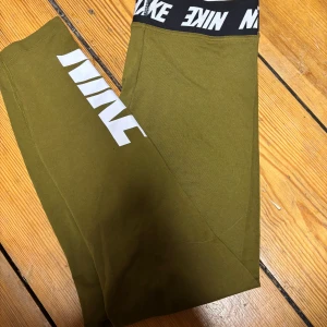 Olivgröna leggings från Nike - Snygga olivgröna leggings från Nike med bred svart resår i midjan och stor vit logga längs benet. Sköna och stretchiga, perfekta för träning eller chill. Tillverkade i mjukt bomullsmaterial med en tight passform som sitter på plats hela dagen.