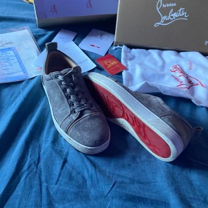 Grå mocka sneakers från Louboutin - Snygga grå sneakers från Christian Louboutin i mocka med klassisk röd sula och rund tå. Skorna har snörning och vita kanter på sulan. Perfekta för dig som vill ha en lyxig och stilren look med en ikonisk touch.