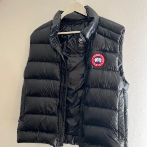 Svart dunväst från Canada Goose - Svart dunväst från Canada Goose med klassisk logga på bröstet. Västen har quiltad design, hög krage och dragkedja framtill. Materialet är glansig polyester och dunfyllning som håller dig varm. Perfekt för lager-på-lager och snygg till hoodie eller tröja. Västen Kommer från johnells Och ja den är äkta 