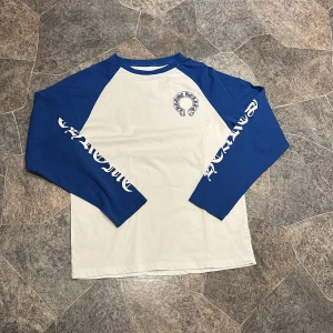 Chrome Hearts långärmad tröja blå/vit - Säljer en långärmad tröja från Chrome Hearts med blå ärmar och vit kropp. Tröjan har tryck på bröstet, stora CH-bokstäver på ryggen och text längs ärmarna. Skön passform och klassisk baseballstil, perfekt för dig som gillar streetwear.