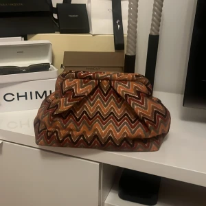 Beck Söndergaard clutch - Snygg kuvertväska från Beck Söndergaard med färgstarkt zigzag-mönster i brunt, orange, beige och vinrött. Väskan har en mjuk form, innerficka med dragkedja och stängs med metallram. 