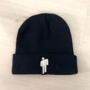En svart Billie Eilish mössa med hennes logga på! - Svart ribbstickad mössa med uppvikt kant och vit broderad logga framtill. Enkel och clean design som passar till det mesta. Perfekt för kalla dagar och streetwear-stil.