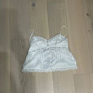 Vit spetsdetaljerad linnetopp - Söt vit linnetopp med tunna axelband och spetsdetaljer längs kanterna. Toppen har dekorativa knappar framtill, broderade detaljer och en romantisk känsla. Perfekt för sommarens varma dagar och passar till både jeans och kjol.
