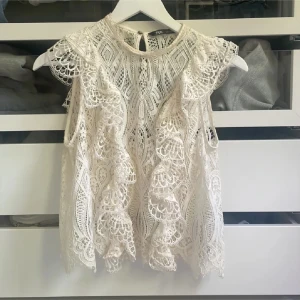 Vit spetsblus från Zara - Superfin vit blus från Zara i spets med vackra volangdetaljer och genombrutet mönster. Ärmlös modell med rund halsringning och knapp i nacken. Perfekt för dig som gillar romantisk och bohemisk stil.