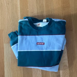 Randig Levi's tröja i blått och grönt - Säljer en snygg randig tröja från Levi's i mörkgrönt och ljusblått med klassisk logga på bröstet. Tröjan har rund hals, långa ärmar och är gjord i mjuk bomull. Perfekt för dig som gillar streetstyle och vill sticka ut lite extra.