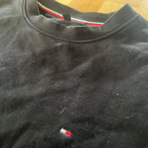 Svart sweatshirt från Tommy Hilfiger - Svart sweatshirt från Tommy Hilfiger med klassisk rund halsringning och diskret broderad logga på bröstet. Tröjan har långa ärmar och ribbade muddar vid ärmslut och nederkant. Perfekt för en clean och stilren look.