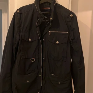 Svart vindjacka från Snoot, storlek L - Svart vindjacka från Snoot med flera fickor framtill, silverfärgade knappar och dragkedjor. Jackan har hög krage, diskret Snoot-logga på ärmen och är tillverkad i ett lätt, slitstarkt syntetmaterial. Perfekt för dig som gillar stilren och funktionell design.