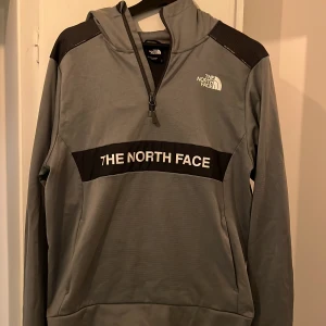 Grå hoodie från The North Face S - Snygg grå hoodie från The North Face med svart panel och vit logga framtill. Tröjan har huva, halv dragkedja och långa ärmar. Perfekt för chill eller träning, med sportig look och bekväm passform.