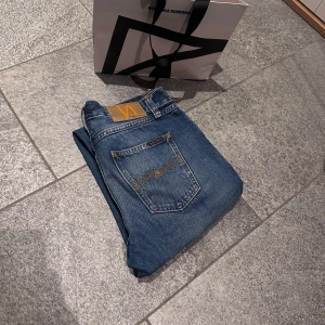 Nudie Jeans blå jeans 29/32 - Snygga blå jeans från Nudie Jeans med klassisk femficksdesign och kontrastsömmar på bakfickorna. Jeansen har en normal passform och är tillverkade i slitstarkt bomullsjeans. Perfekta för dig som gillar stilrena och tidlösa jeans.