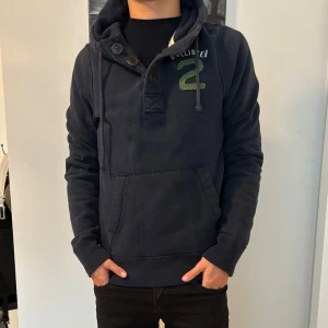 Mörkblå hoodie från Hollister - Snygg mörkblå hoodie från Hollister med grönt tryck och broderad siffra på bröstet. Tröjan har huva med snörning, knappar vid halsen. Mjuk och skön i bomullsmaterial, perfekt för chill dagar. Passformen är normal, hör av vid funderar!