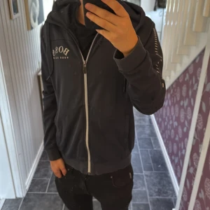 Mörkgrå hoodie från Hugo Boss - Snygg mörkgrå hoodie från Hugo Boss med vit dragkedja och logga på bröstet. Tröjan har huva, långa ärmar med vita detaljer och två fickor framtill. Perfekt för dig som gillar stilrena streetwear-plagg med premiumkänsla. Tydliga defekter på armbågarna och där framme vid dragkedjan därför billigt pris. Sitter bra på mig som är 170