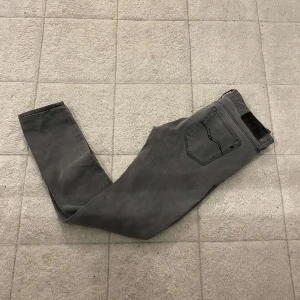 Replay grå jeans slim fit - Snygga grå jeans från Replay, modell Anbass. Slim fit med klassisk femficksdesign och svarta detaljer på bakfickan. Jeansen har väldigt små slitningar vid låret som knappt syns. Jeansen har normal midja och är tillverkade i mjukt bomullsmaterial med stretch för extra komfort.