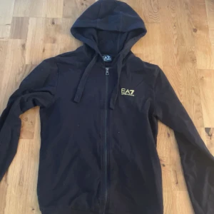 Xs/S Svart hoodie från EA7 Emporio Armani - Svart hoodie från EA7 Emporio Armani med gul logga på bröstet. Tröjan har dragkedja framtill, huva med snörning och långa ärmar. Perfekt för en chill och sportig look.