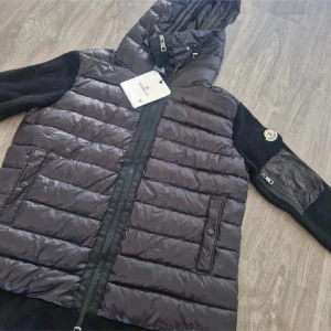 Moncler cardigan - Svart dunväst från Moncler med quiltad front och huva. Västen har stickade svarta ärmar med Moncler-logga på vänster arm och en ficka med dragkedja. Framtill finns två fickor med knappar och en hel dragkedja. Snygg och sportig design, perfekt för kyliga dagar.