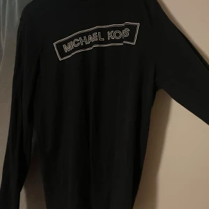 Svart långärmad tröja Michael Kors - Svart långärmad tröja från Michael Kors med vit logga framtill. Tröjan är i mjuk bomulls- och polyestermix, perfekt för en clean och stilren look. Enkel passform och klassisk rund halsringning.